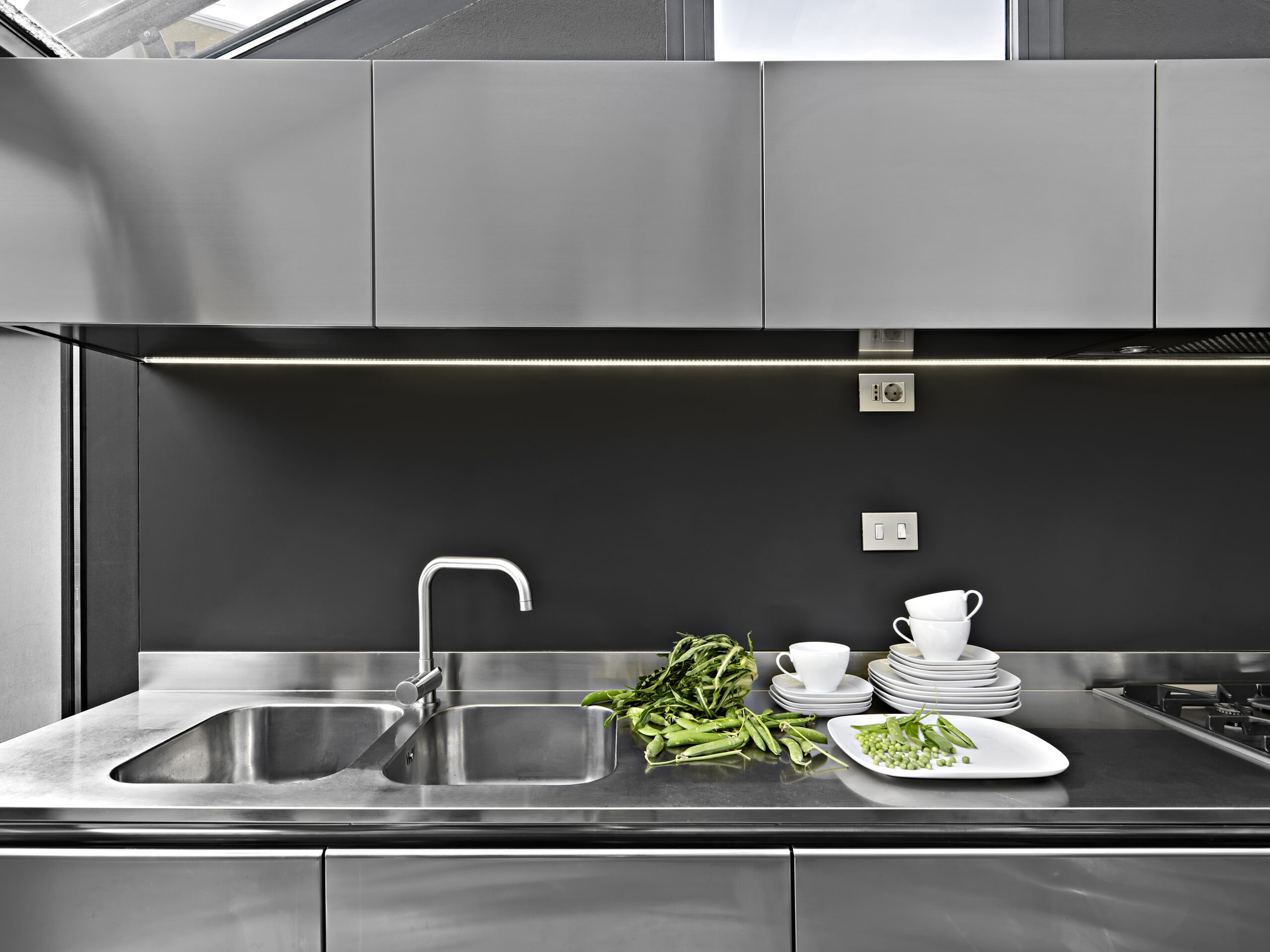 modern interior of the kitchen in the foreground the steel sink with integrated sink near which there are tea cups and fresh vegetables, the wall units are made of steel